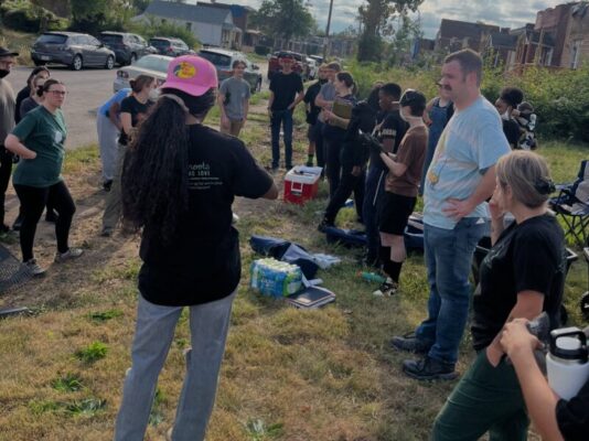 A woman wearing a pink hat stands facing a large group of volunteers gathered on a grassy curb alongside a residential street. She appears to be giving instructions or a briefing before a neighborhood cleanup event.