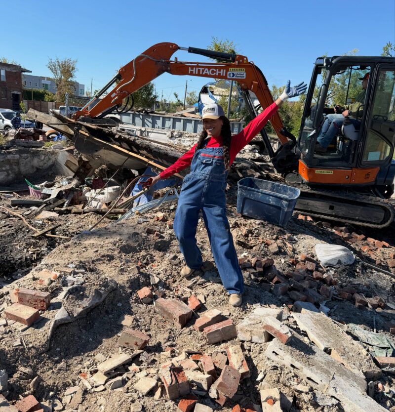 A smiling woman in denim overalls and a St. Louis cap poses with her arms outstretched in front of a small orange Hitachi excavator. The machine and a large pile of housing debris fill the active demolition site behind her.