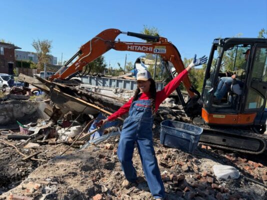 A smiling woman in denim overalls and a St. Louis cap poses with her arms outstretched in front of a small orange Hitachi excavator. The machine and a large pile of housing debris fill the active demolition site behind her.