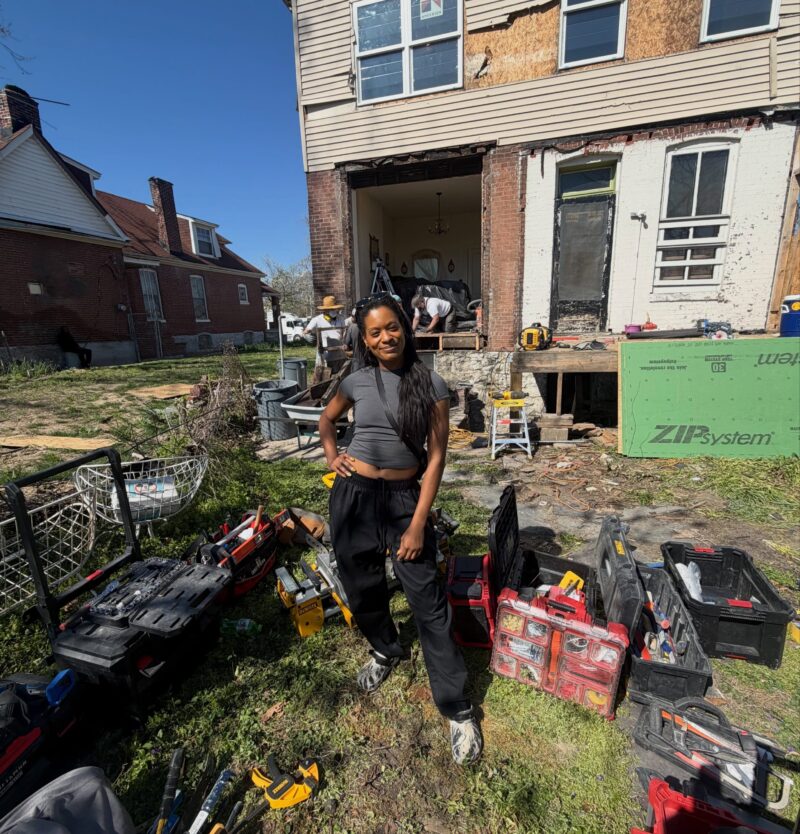 A smiling woman poses in a grassy yard surrounded by several open toolboxes filled with construction tools. In the background, workers are renovating the exterior of a brick house using green ZIP System sheathing.