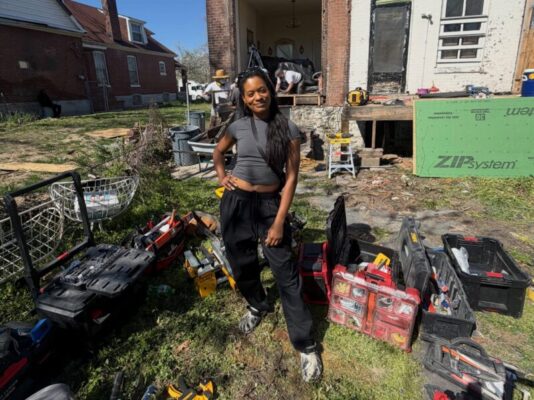 A smiling woman poses in a grassy yard surrounded by several open toolboxes filled with construction tools. In the background, workers are renovating the exterior of a brick house using green ZIP System sheathing.